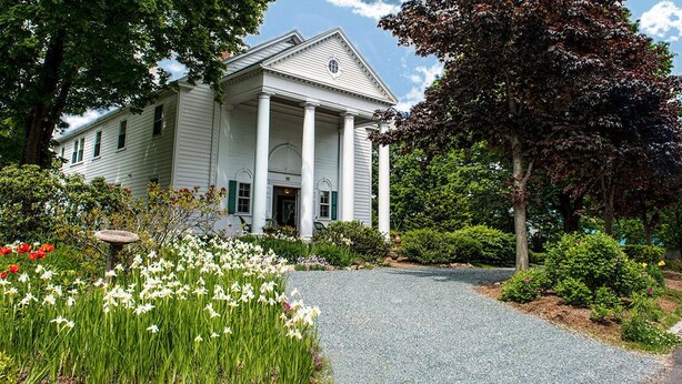 A white building with columns, surrounded by a gravel driveway and lush gardens.