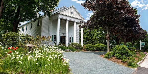 A white building with columns, surrounded by a gravel driveway and lush gardens.