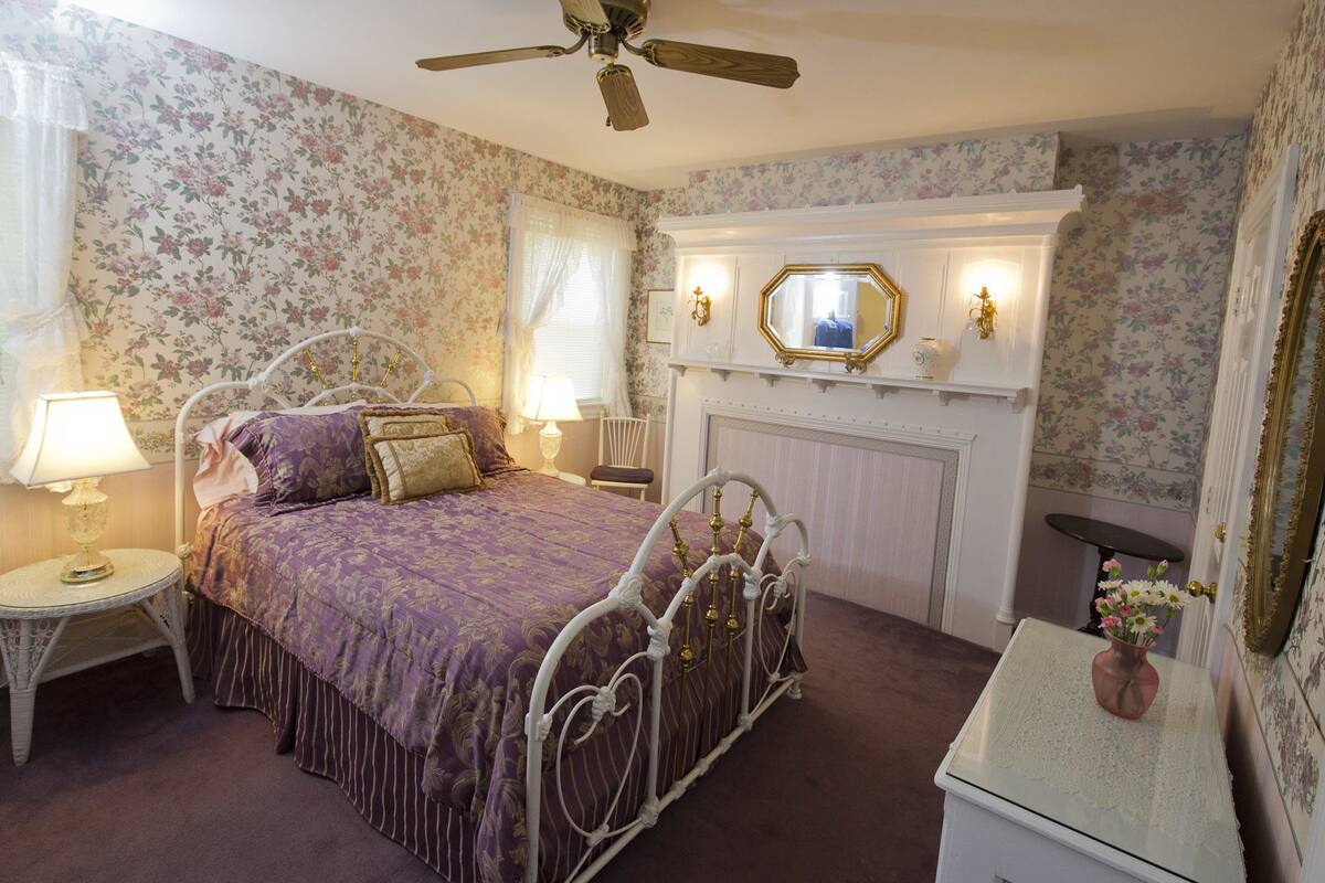 A bedroom with a four-poster bed, a ceiling fan, a fireplace, and floral wallpaper.