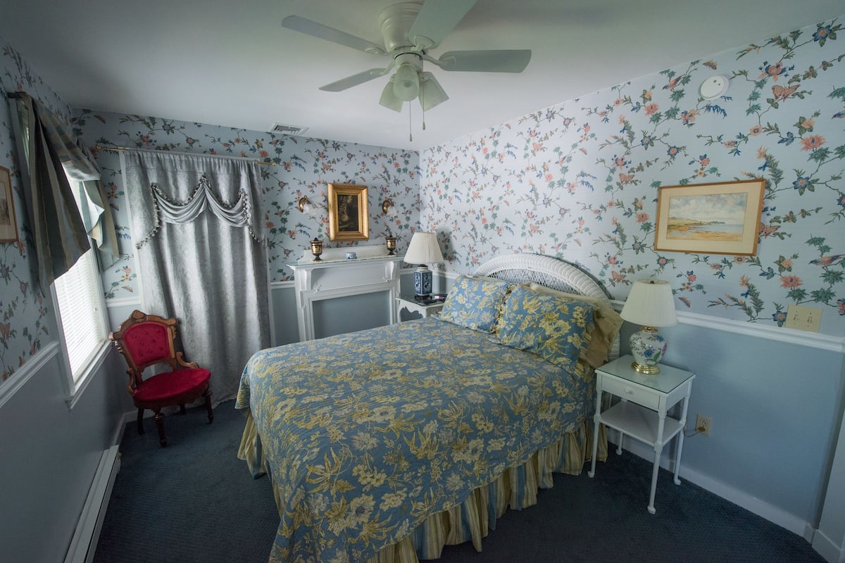 A bedroom with a bed, a chair, a nightstand, a ceiling fan, and floral wallpaper.