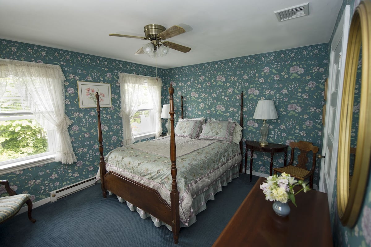 A four-poster bed with floral bedding, a ceiling fan, and a window with lace curtains.