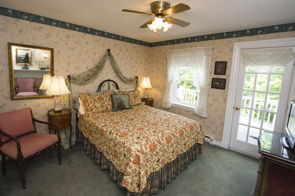 A bedroom with a bed, two bedside tables, a chair, a ceiling fan, a window with curtains, and a door.