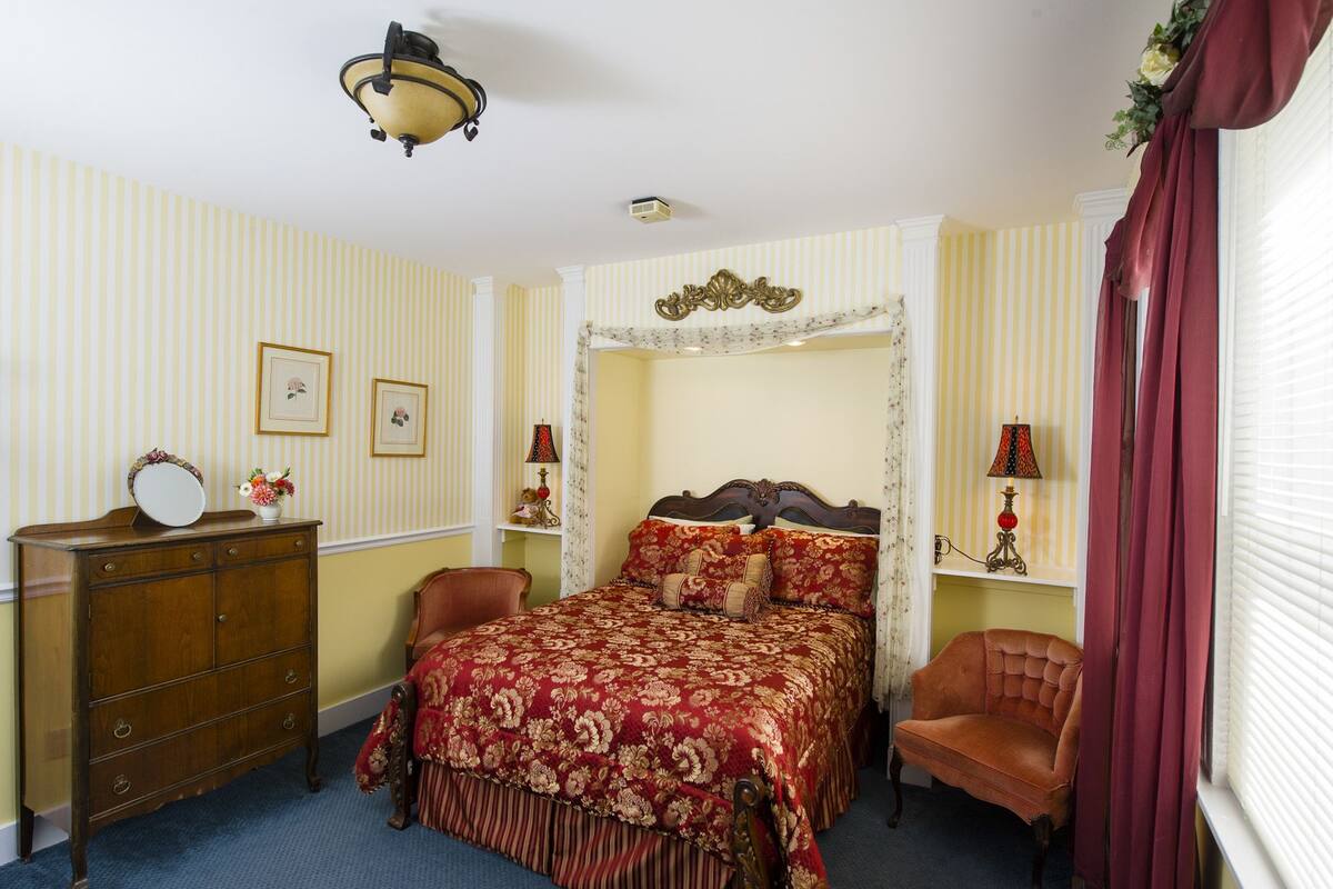 A bedroom with a four-poster bed, a wooden dresser, a chair, and a window with curtains.