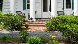 A white building with green shutters, a brick entrance, and a garden with potted plants and yellow flowers.