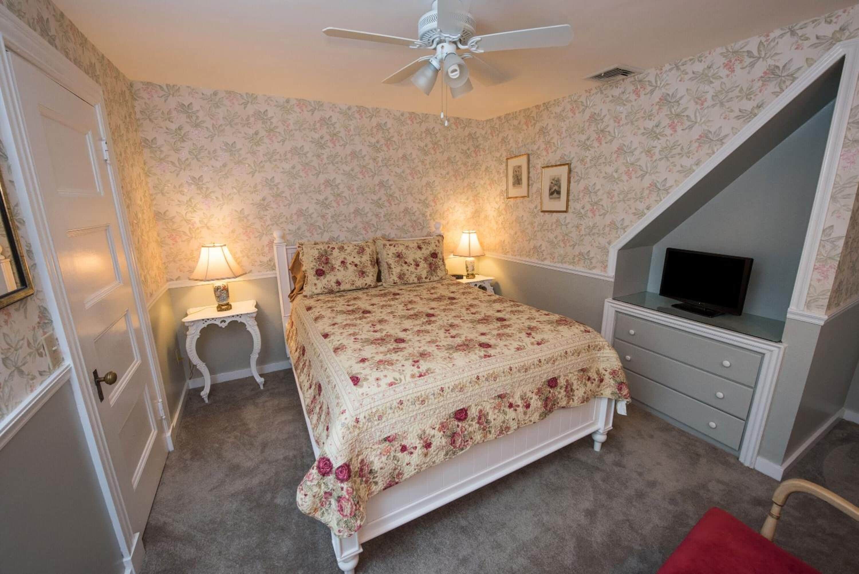 A bedroom with a bed, two bedside lamps, a television on a stand, and a ceiling fan.