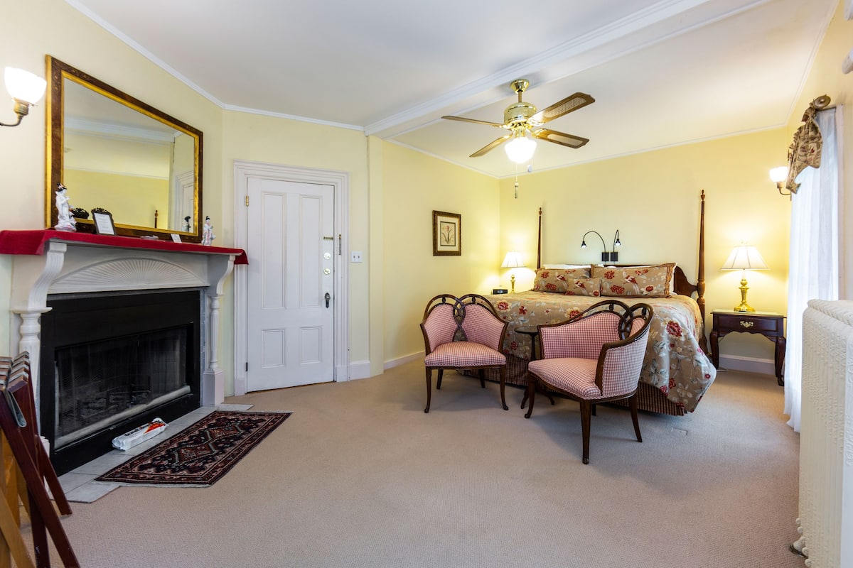 A bedroom with a fireplace, a ceiling fan, a bed with floral bedding, two chairs, and a nightstand with lamps.