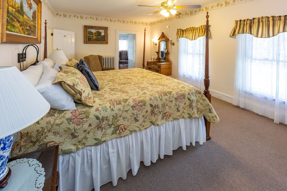 A bedroom with a four-poster bed, a ceiling fan, a radiator, and a view of the hallway.