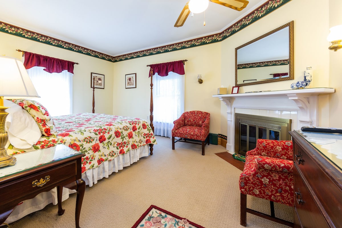 A bedroom with a bed, a chair, a dresser, a mirror, and a fireplace.