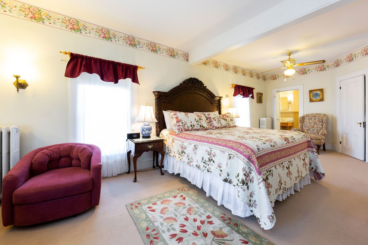 A bedroom with a large bed, a red armchair, a nightstand, and a floral-patterned rug.