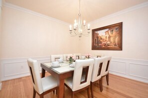 A dining room with a wooden table set for four, white chairs, a chandelier, and a framed painting on the wall.