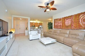 A living room with a sofa, a coffee table, and a ceiling fan. There is a kitchen in the background with cabinets and a microwave.