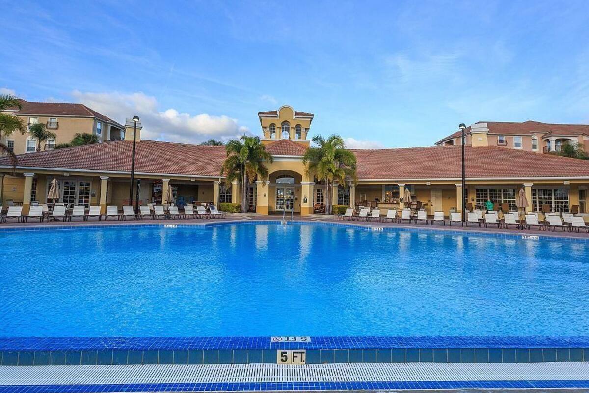 A large swimming pool with lounge chairs and umbrellas, surrounded by buildings with red-tiled roofs and a central tower.