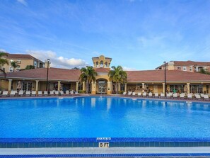 A large swimming pool with lounge chairs and umbrellas, surrounded by buildings with red-tiled roofs and a central tower.