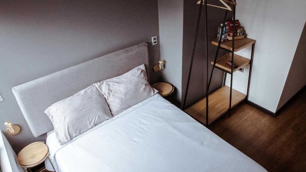 Un lit bien fait, avec du linge de lit blanc, une tête de lit grise et une table de chevet en bois, à côté de laquelle se trouve une petite table ronde.
