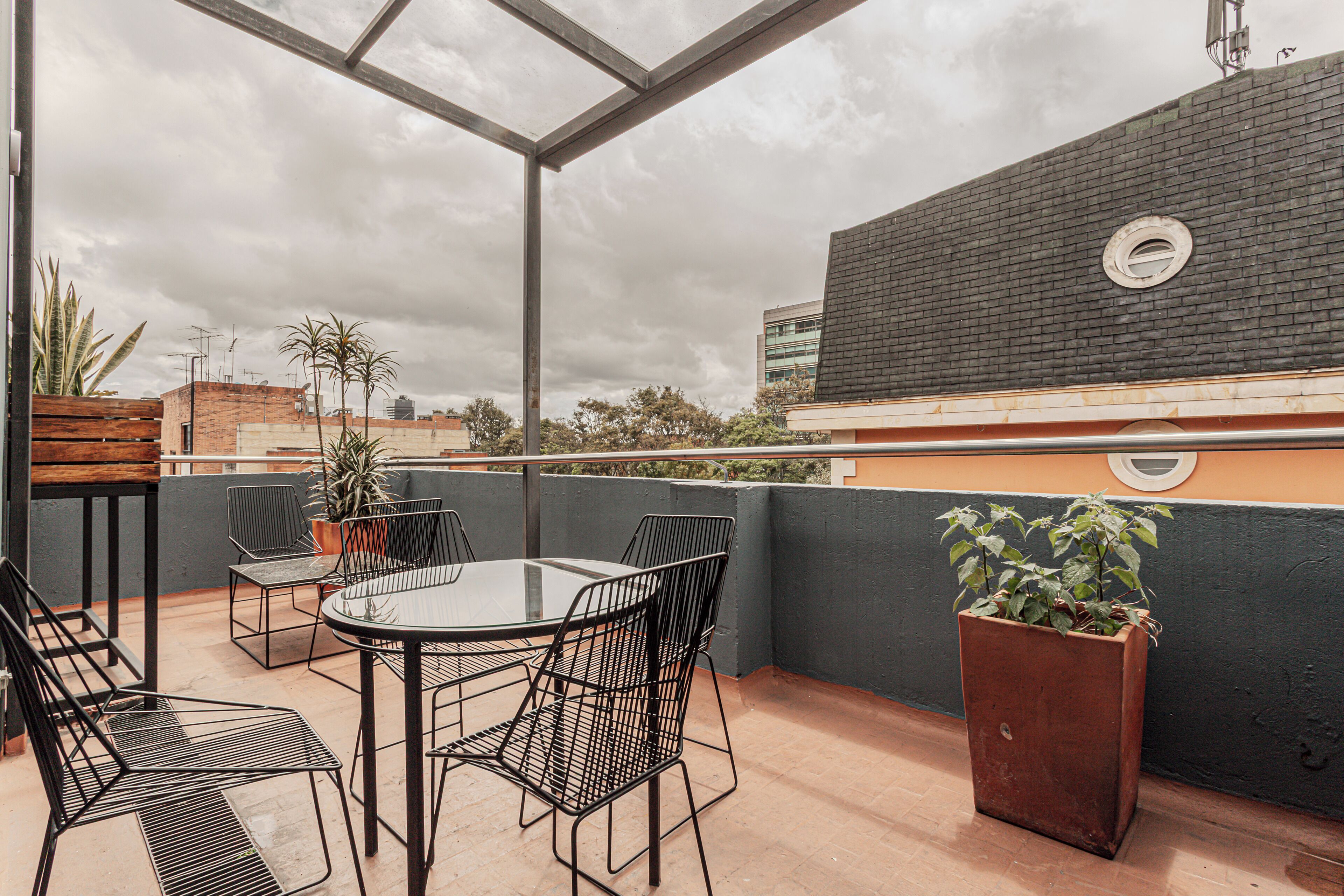 Una terraza en la azotea con una mesa de vidrio y sillas de metal, rodeada de plantas en macetas y una jardinera grande.