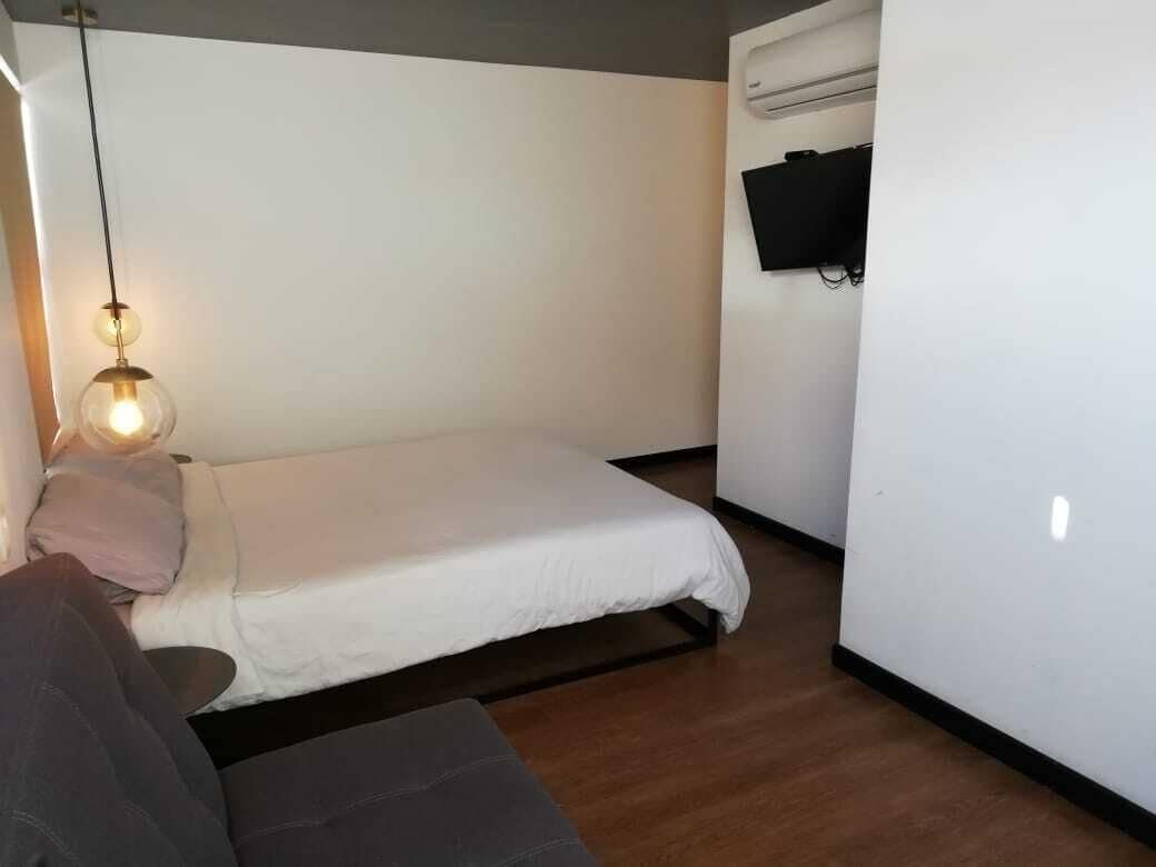 A small, single-bed room with a wooden floor, a white wall, a mounted TV, and a hanging light fixture.