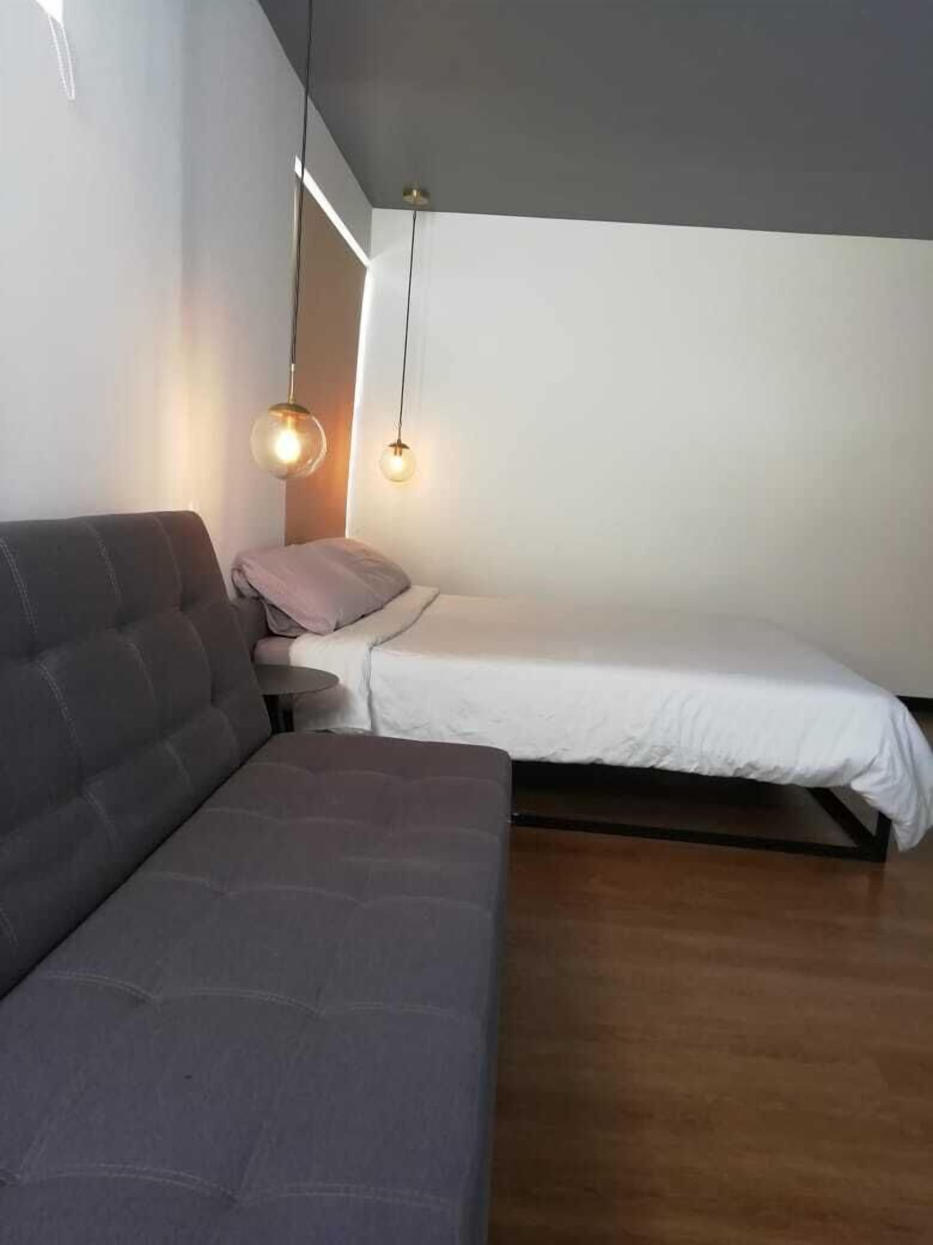 Une chambre à coucher moderne avec un canapé gris, une petite table de chevet et un lit simple doté d’un matelas blanc.