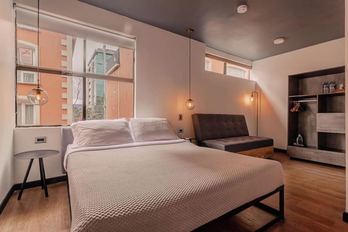Un dormitorio moderno con una cama grande, una mesita, una silla, un armario y una ventana con vistas a edificios.
