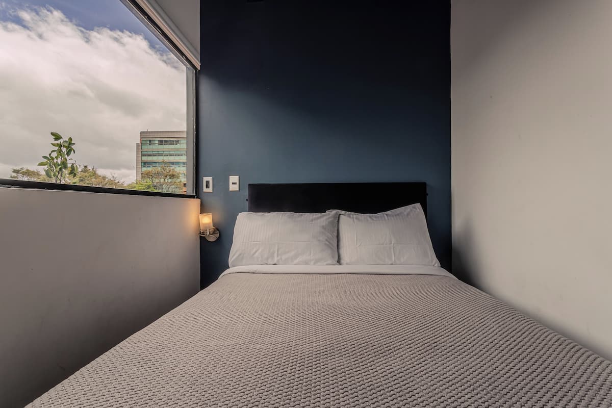 Una cama con ropa de cama blanca, cabecera oscura y ventana con vistas a edificios y árboles.