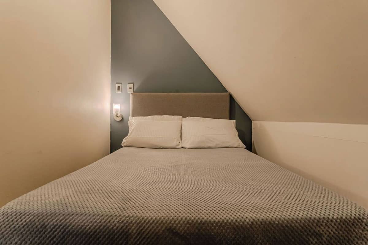 Una habitación pequeña y estrecha con una cama, una luz fijada en la pared y un techo inclinado.