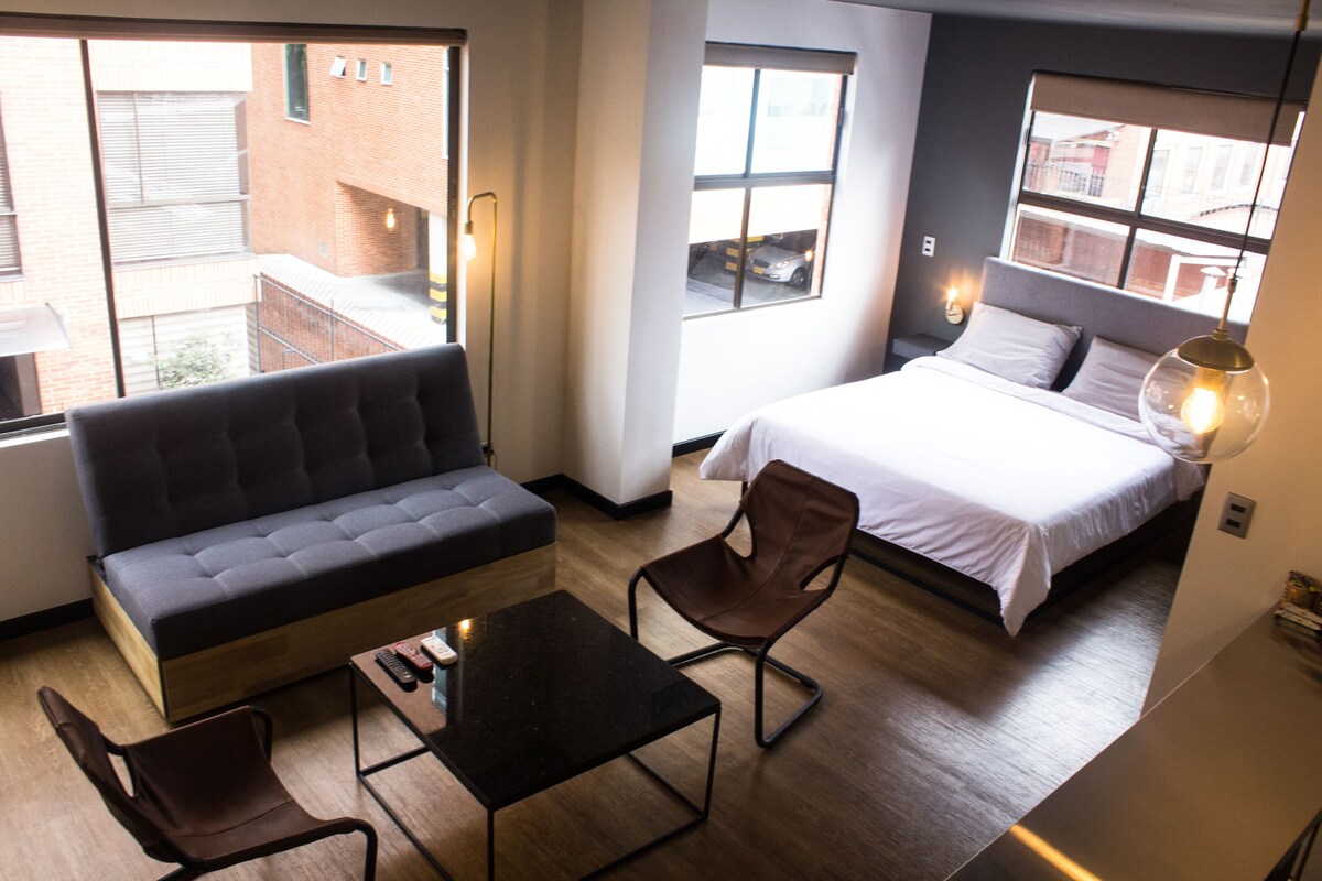 A modern hotel room with a sofa, two chairs, a small table, a bed, and large windows offering a view of the city.