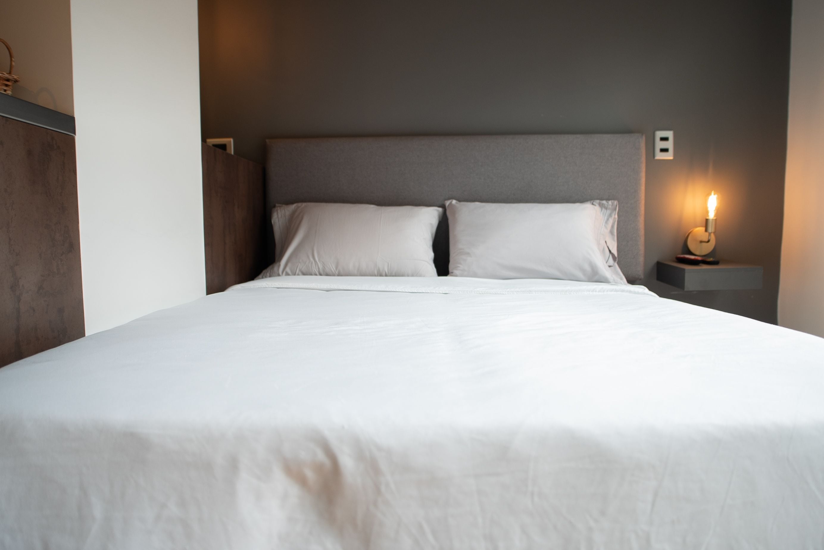 A neatly made bed with white linens, a grey headboard, and a bedside table with a lit candle.