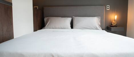 A neatly made bed with white linens, a grey headboard, and a bedside table with a lit candle.