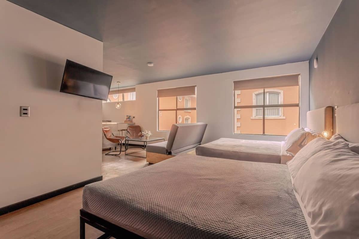 Habitación de hotel moderna con una cama grande, una cama más pequeña, un televisor montado en la pared y vistas de la ciudad a través de grandes ventanales.
