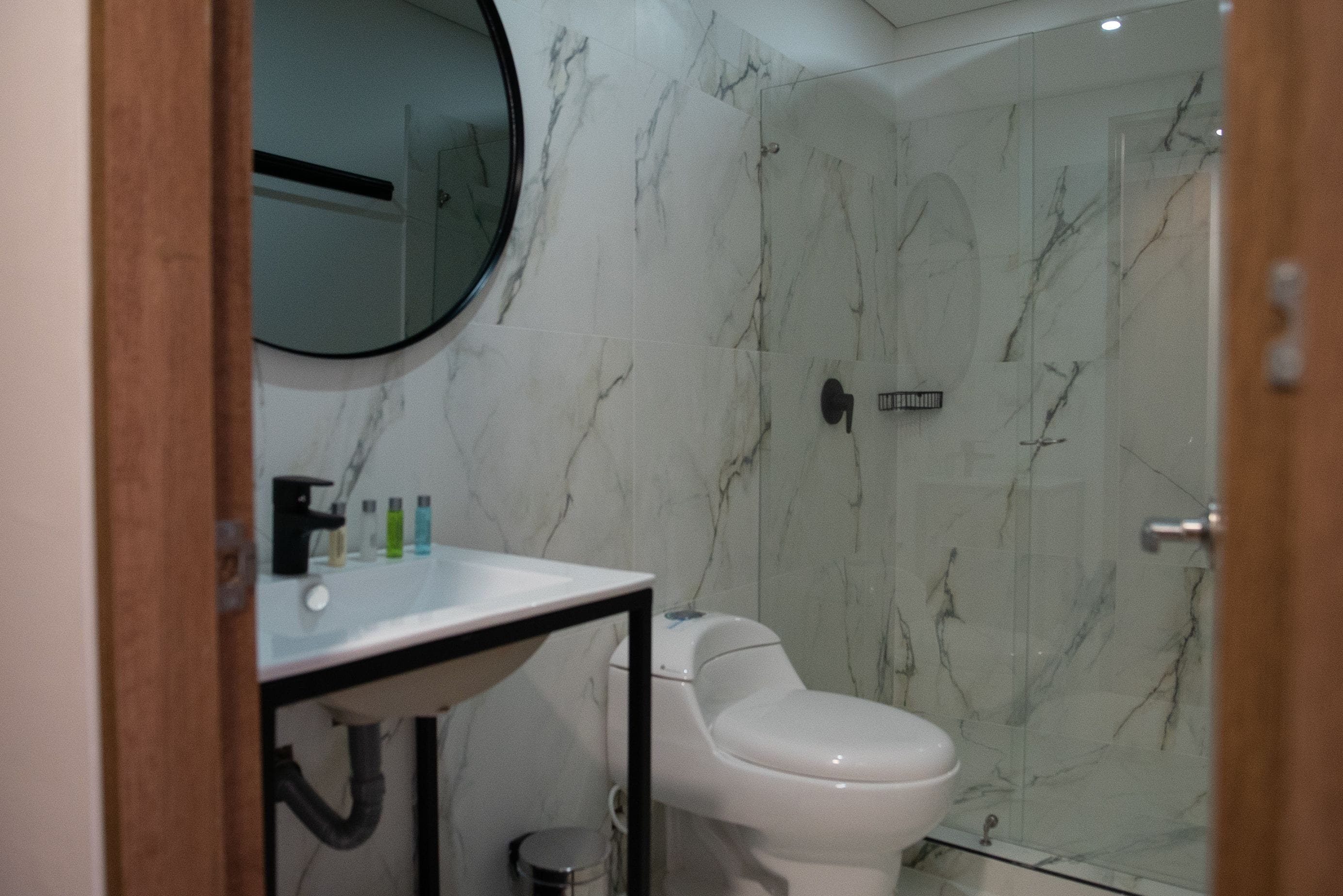 A modern bathroom with a marble wall, a round mirror, a toilet, and a sink with toiletries.