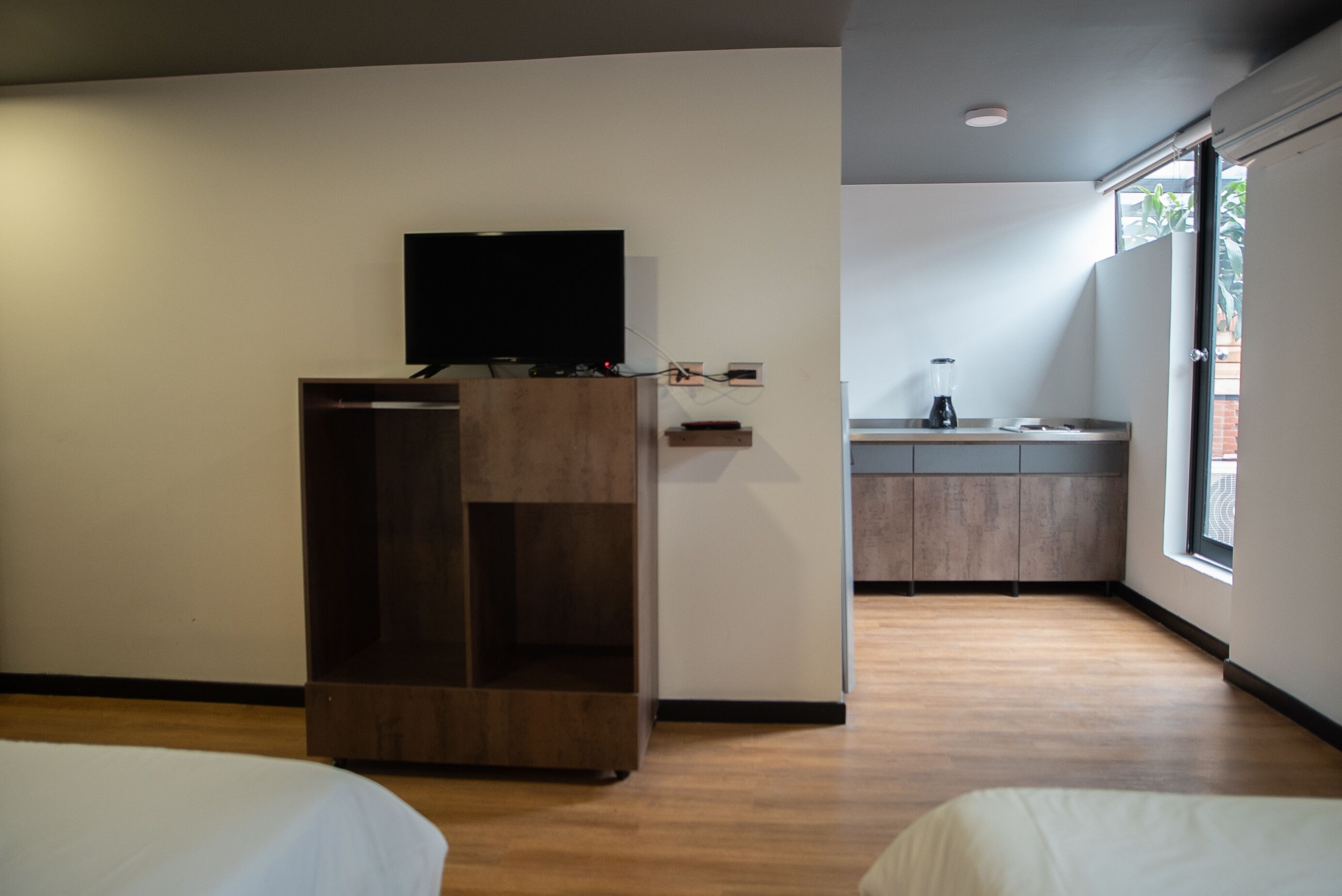 A hotel room with a bed, a TV mounted on a wooden cabinet, a kitchenette with a blender, and a large window.