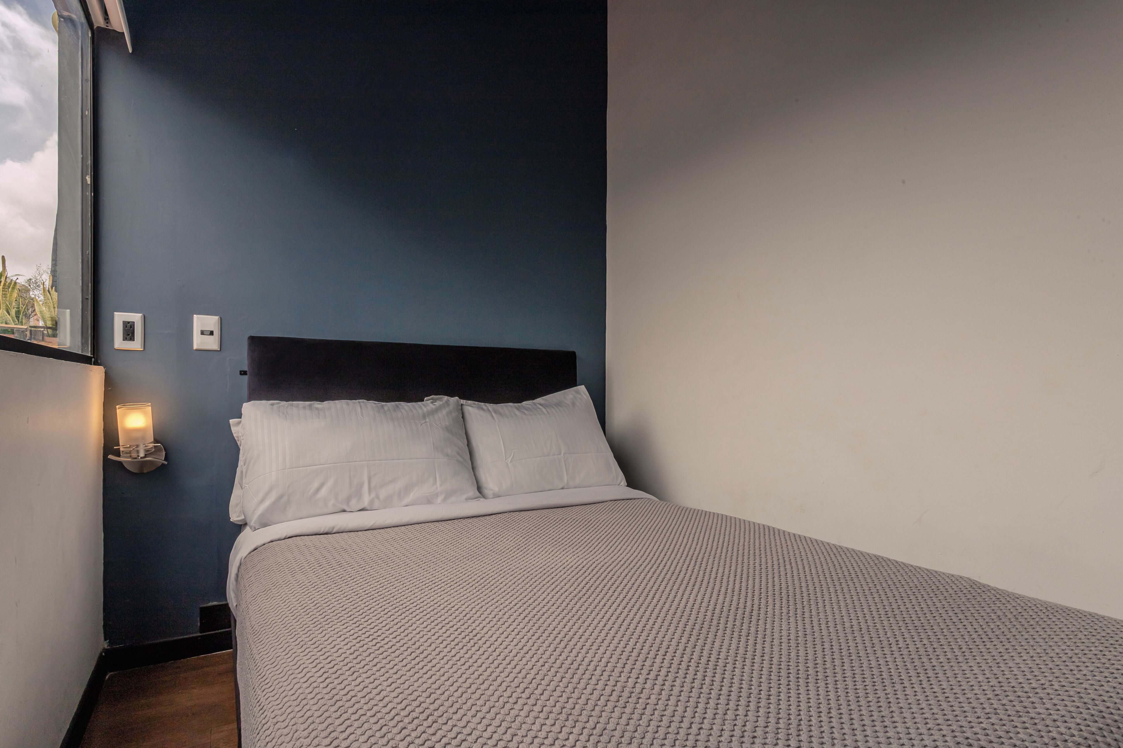 Een slaapkamer met een donkerblauwe wand, een wit bed met een geribbelde deken, een zwart hoofdeinde en een wandlamp.