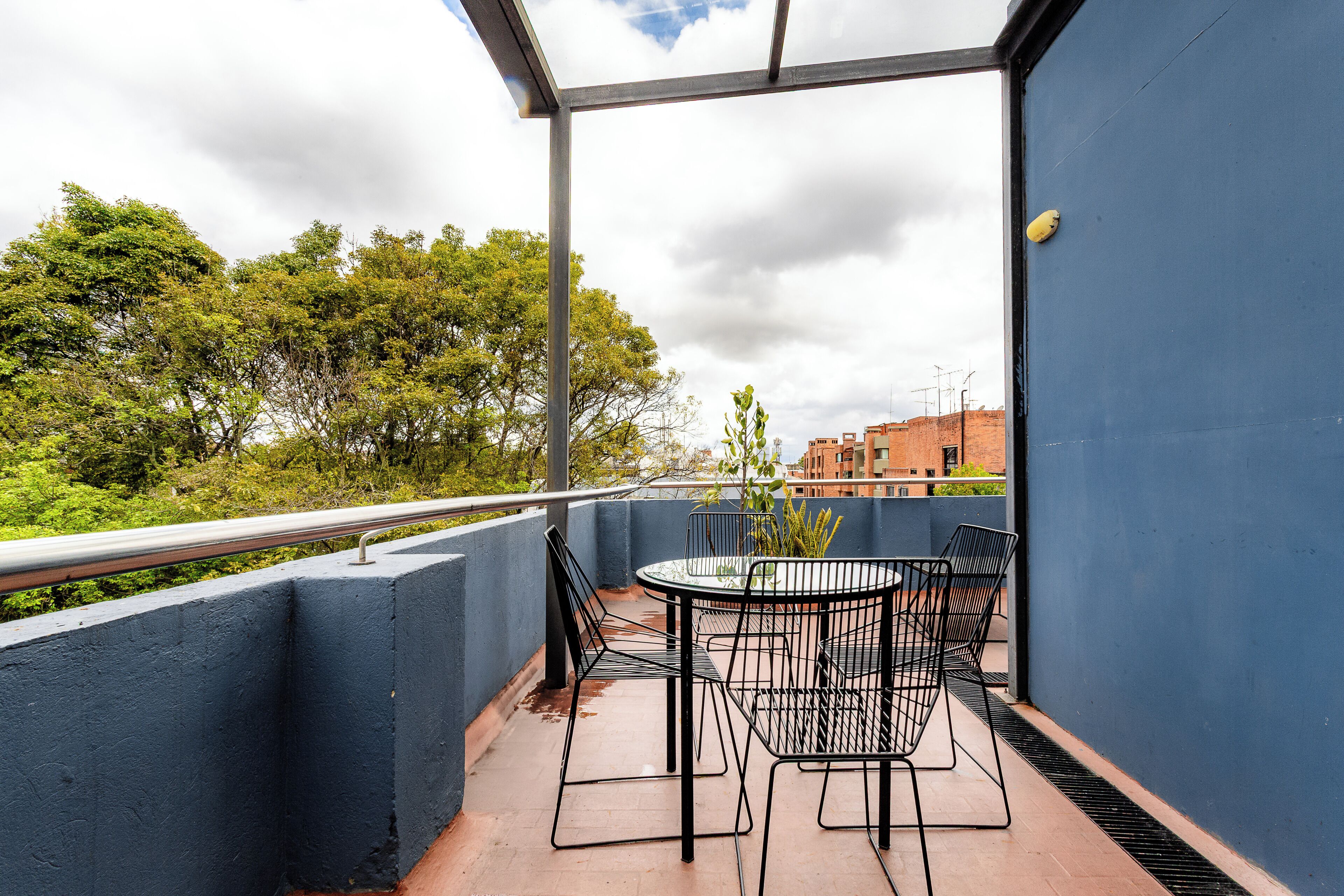 A balcony with a round table and chairs, a view of trees and buildings, and a blue wall.