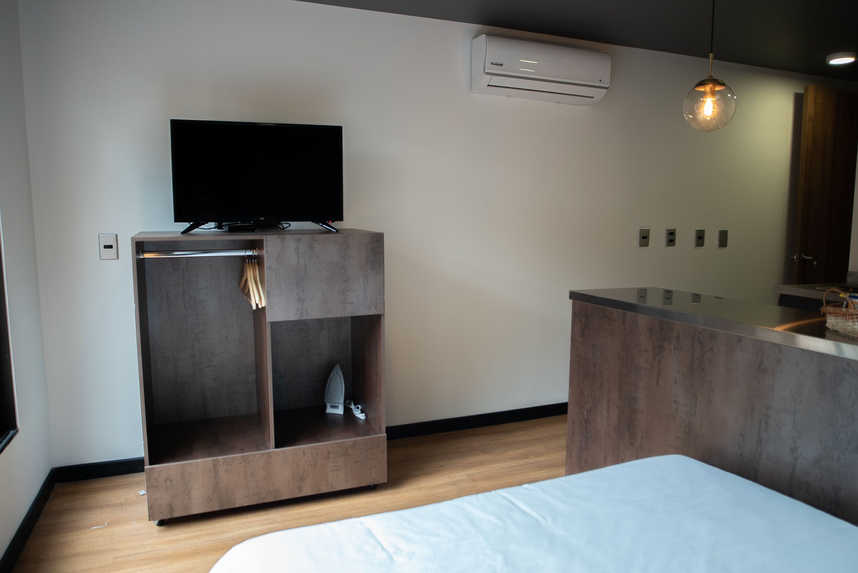 Moderni hotellihuone, jossa on seinälle asennettu taulutelevisio, puulattia, valkoinen sänky ja pieni oleskelualue, jossa on tuoli ja pöytä.