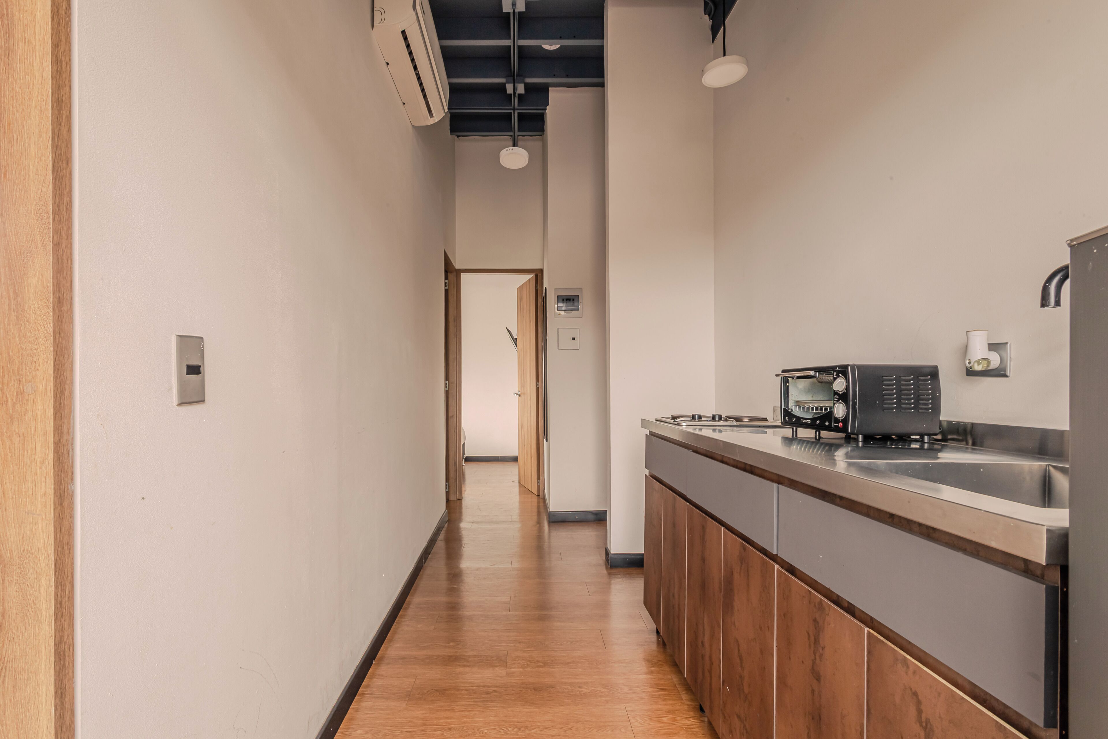 A narrow hallway with a wooden floor, a kitchen area with a microwave, toaster, and sink, and a door leading to another room.