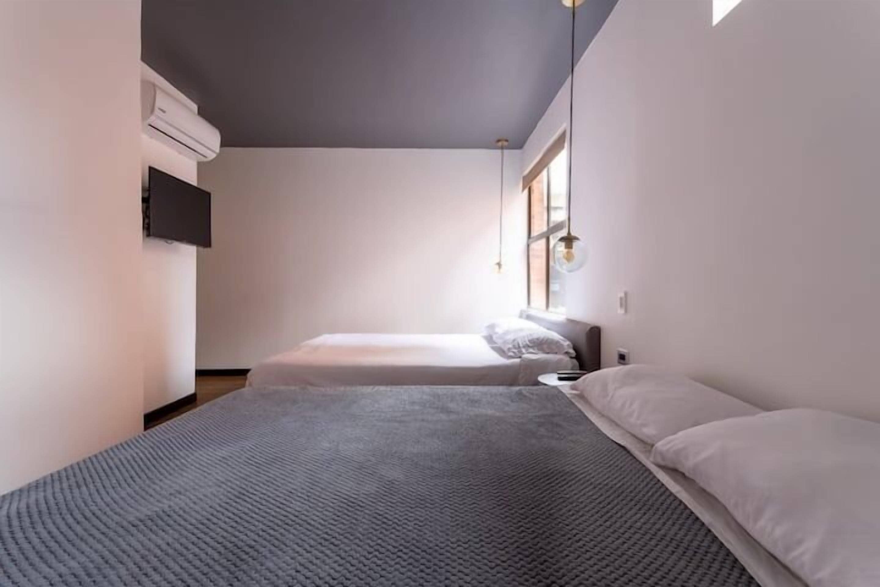 A minimalist bedroom with two beds, a flat-screen TV, and a window providing natural light.