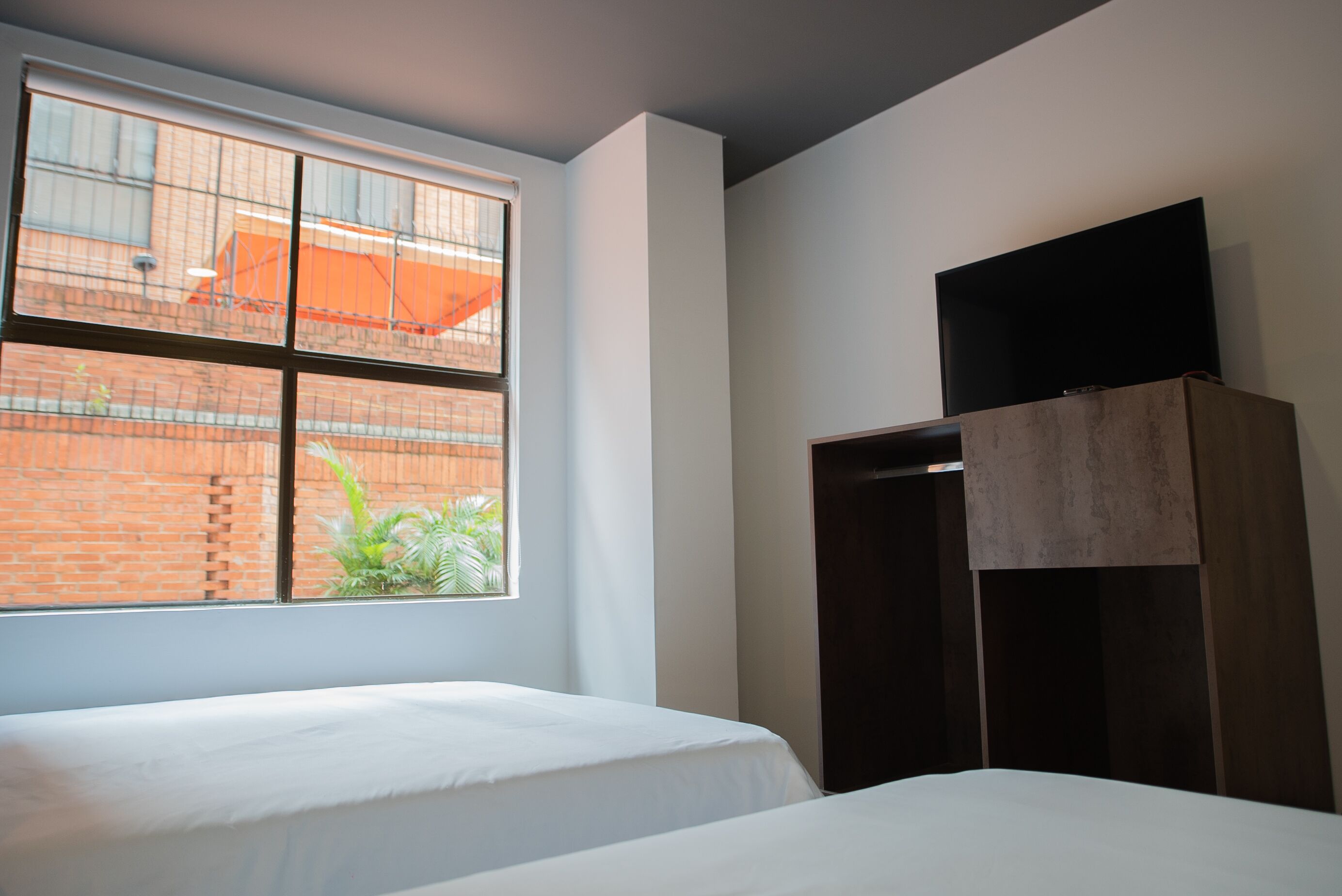 A hotel room with a large window, a bed, a wardrobe, and a flat-screen TV mounted on the wall.