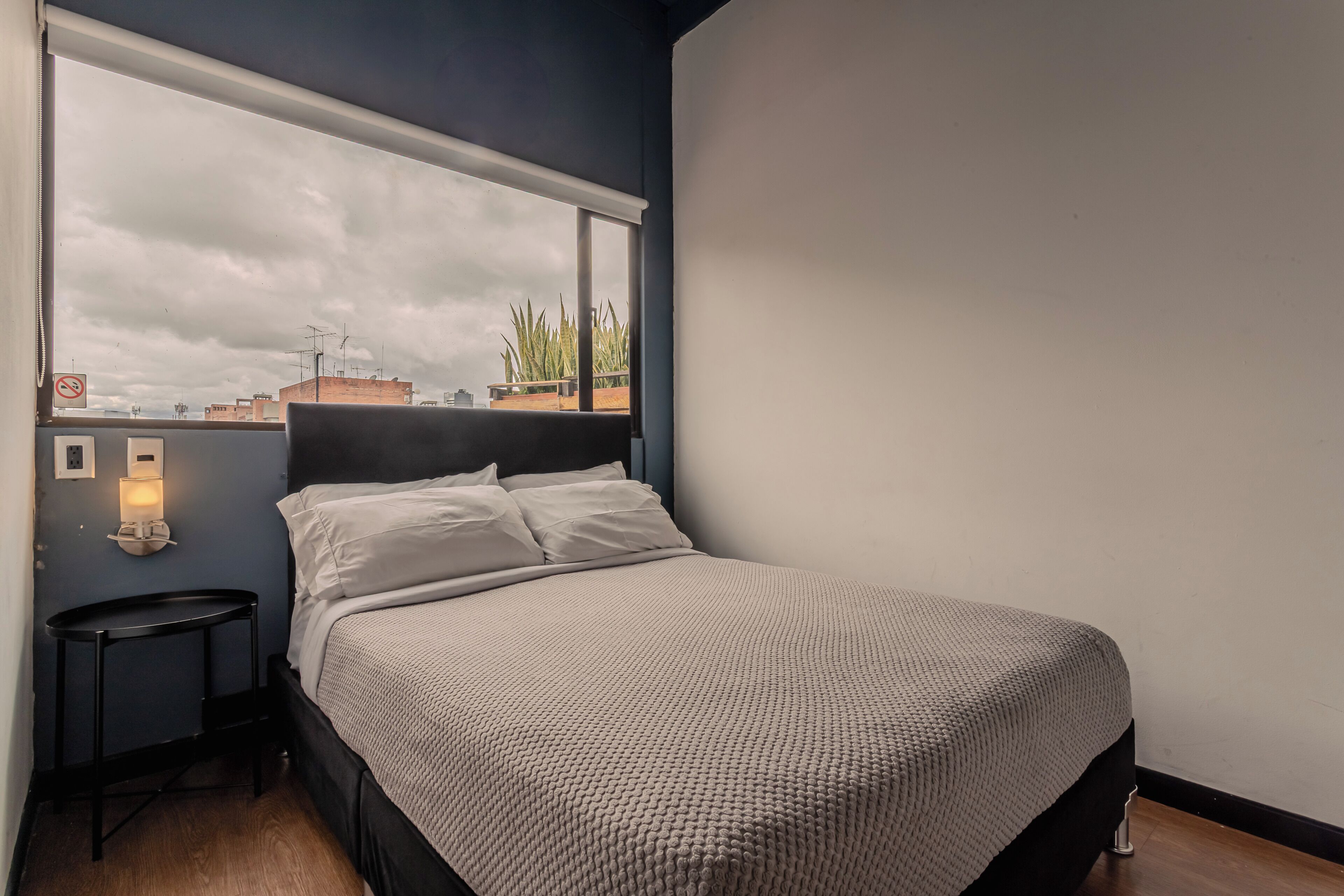 A bedroom with a bed, a nightstand, a window with a view of buildings, and a wall-mounted light.