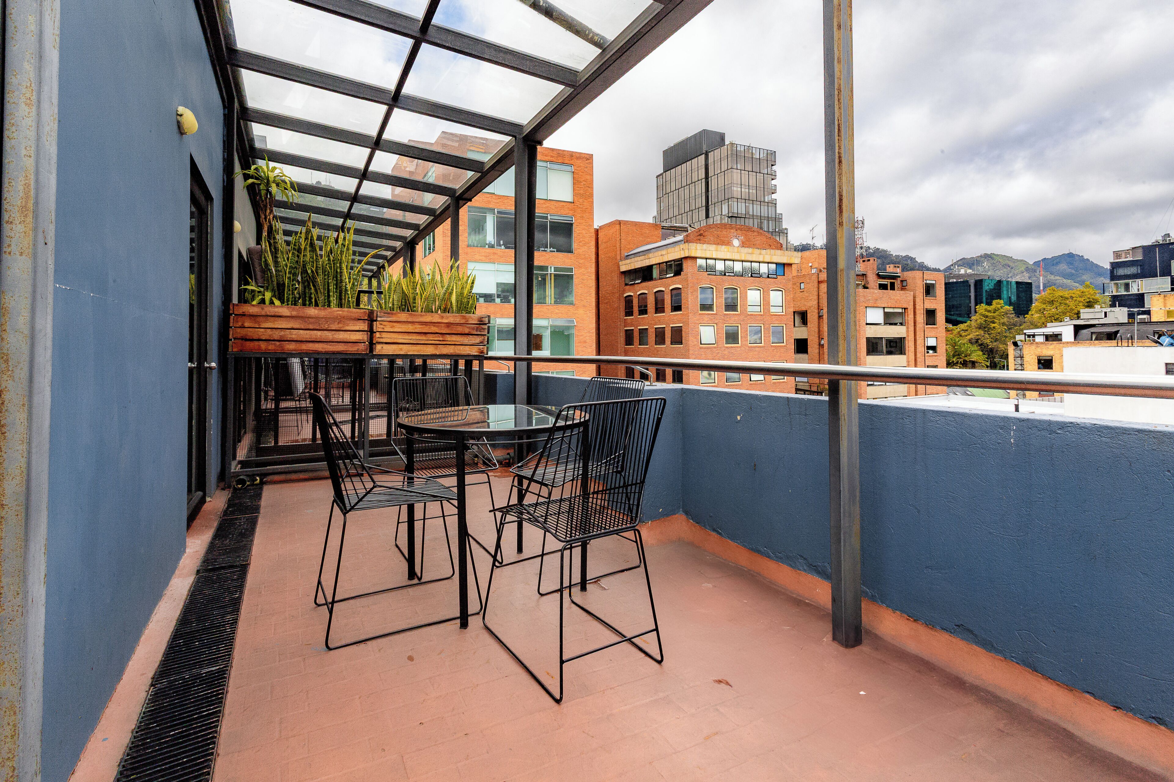 A balcony with a glass enclosure, a wooden planter with plants, a round table, and two chairs.