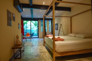A four-poster bed with a white mattress, a wooden bench with a statue, and a view of greenery through large windows. - La Jolla Hotel (Cóbano)