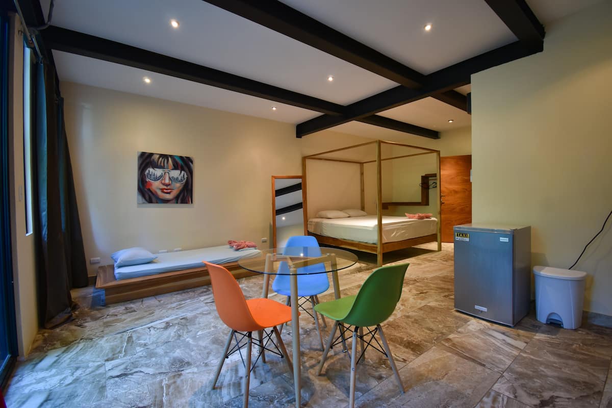 A compact room with a bed, a small table with chairs, a refrigerator, and a painting on the wall.