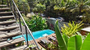 A pool area with wooden steps leading down to a pool, surrounded by lush greenery and a view of a street with cars. - La Jolla Hotel (Cóbano)