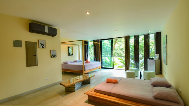 A modern hotel room with a large bed, a smaller bed, a bench, and a desk. There are large windows with a view of greenery outside. - La Jolla Hotel (Cóbano)