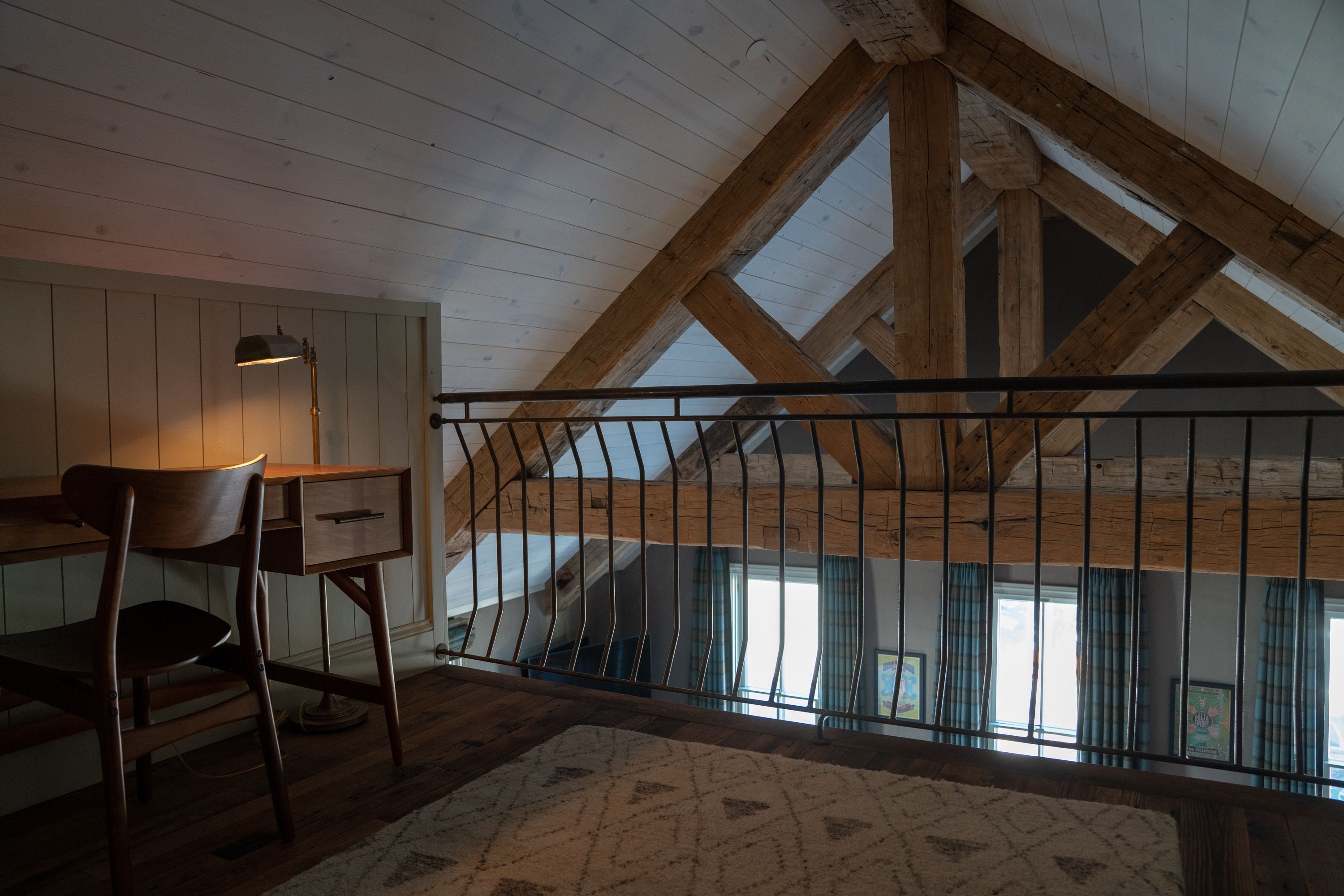 A loft bedroom with a sloped ceiling, wooden beams, a desk with a chair, and a view through a window with curtains.