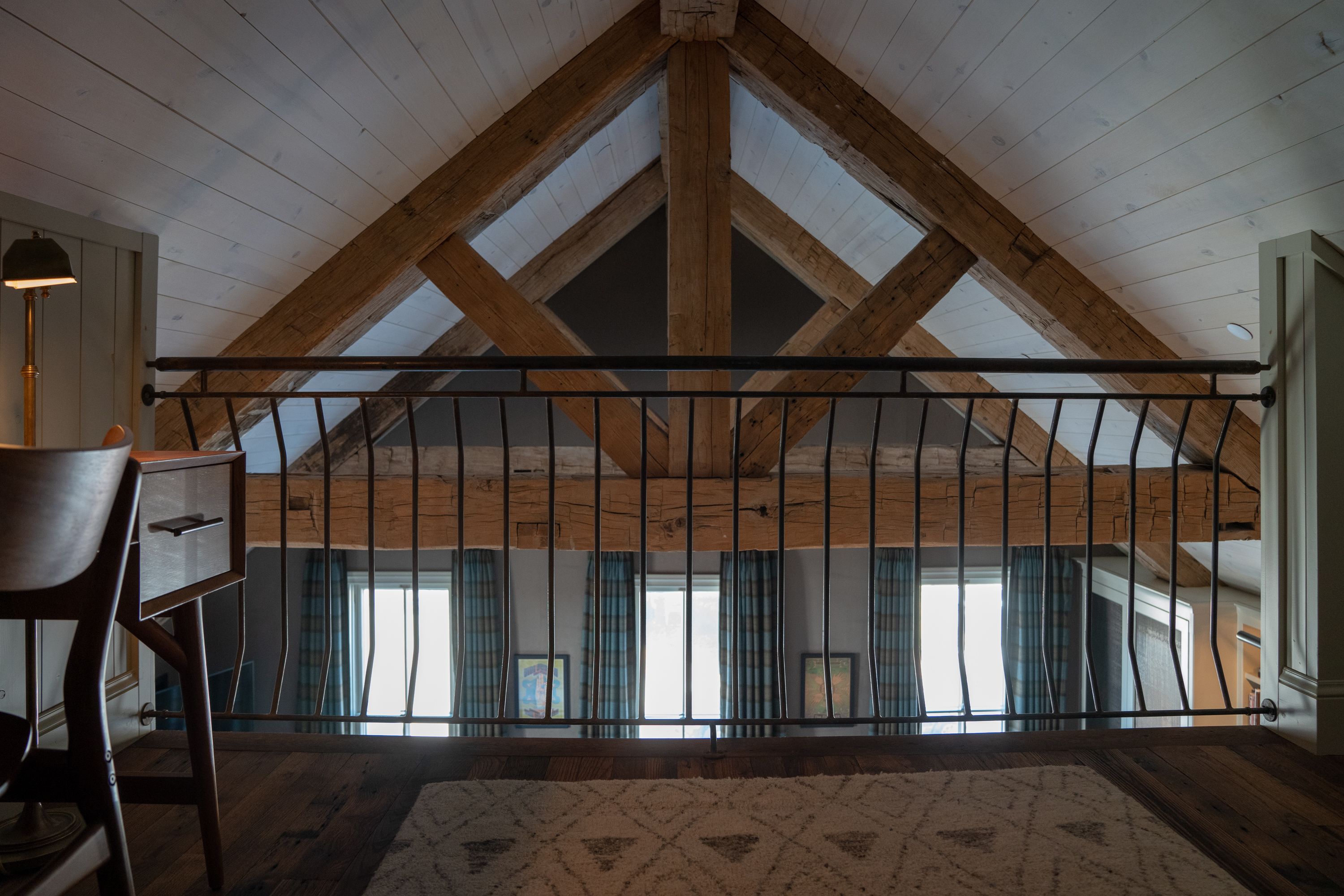 Ein Zimmer mit hohen Decken, freiliegenden Holzbalken, einem großen Fenster mit Vorhängen und einem gemusterten Teppich auf dem Boden.