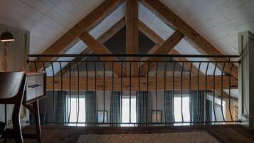 A high-ceilinged room with exposed wooden beams, a large window with curtains, and a patterned rug on the floor.