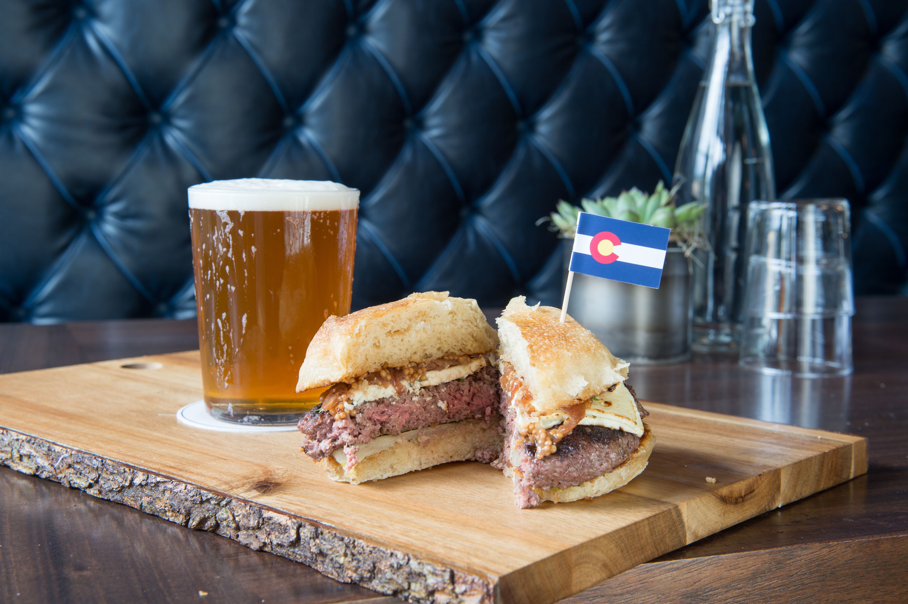 A sandwich cut in half on a wooden board, a glass of beer, and a bottle with a flag.