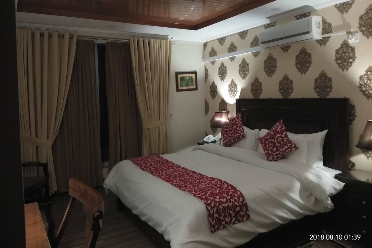 Una habitación de hotel con cama, cortinas, aire acondicionado, lámpara y mesita de noche.