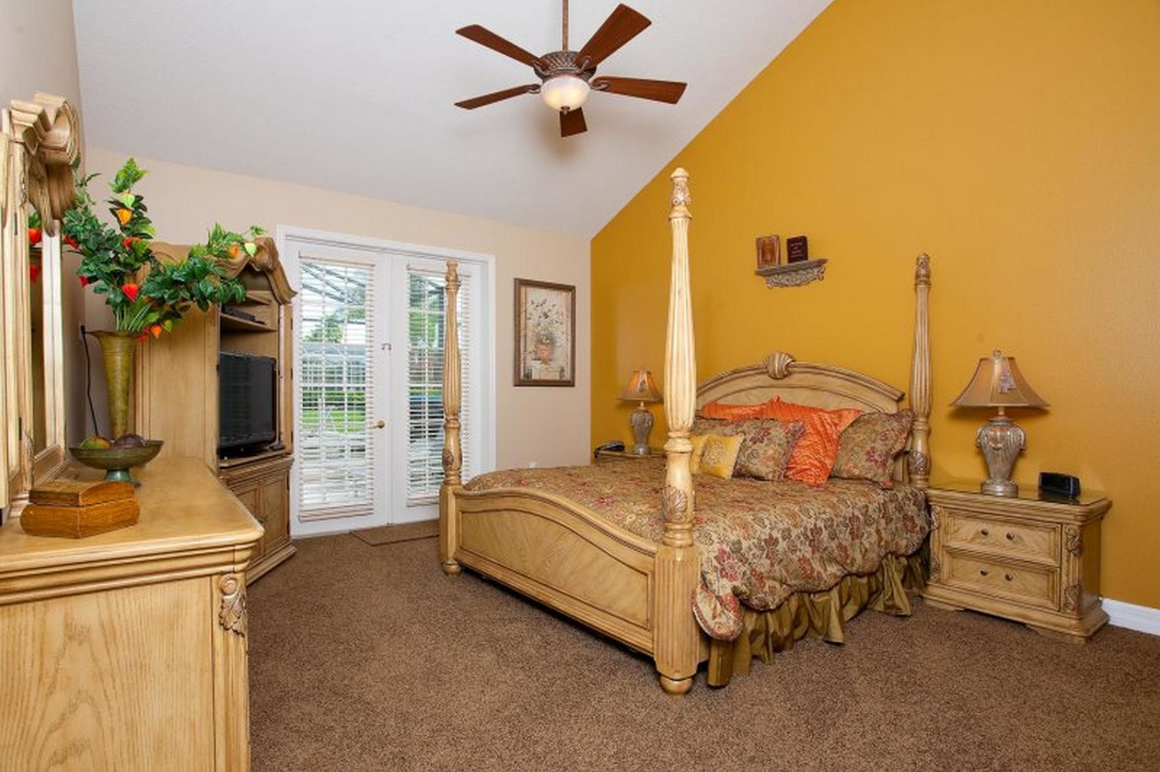 A bedroom with a four-poster bed, a ceiling fan, a nightstand, a lamp, a dresser, a mirror, a plant, and a window with blinds.