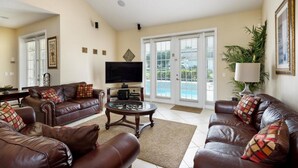 A living room with leather sofas, a coffee table, a flat-screen TV, and a large window with a view of a pool. - Formosa Gardens Homes by Atlas VH (Kissimmee)