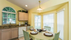 A dining area with a table set for four, a kitchen with a window, and a ceiling light fixture. - Indian Creek Homes by Atlas VH (Kissimmee)